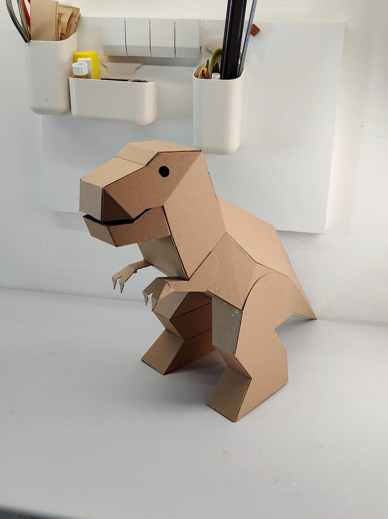 May include: A geometric dinosaur figure constructed from light brown cardboard. The sculpture features a blocky, angular design with a large head, small arms, and a thick tail, showcasing a clean, folded paper aesthetic with a simple black dot eye.
