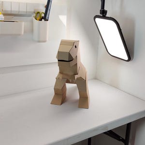 May include: A three-dimensional dinosaur figure constructed from light brown cardboard. The sculpture features a blocky, geometric design with a distinct head, a wide mouth, two short front limbs, and two sturdy back legs, all assembled from flat panels.