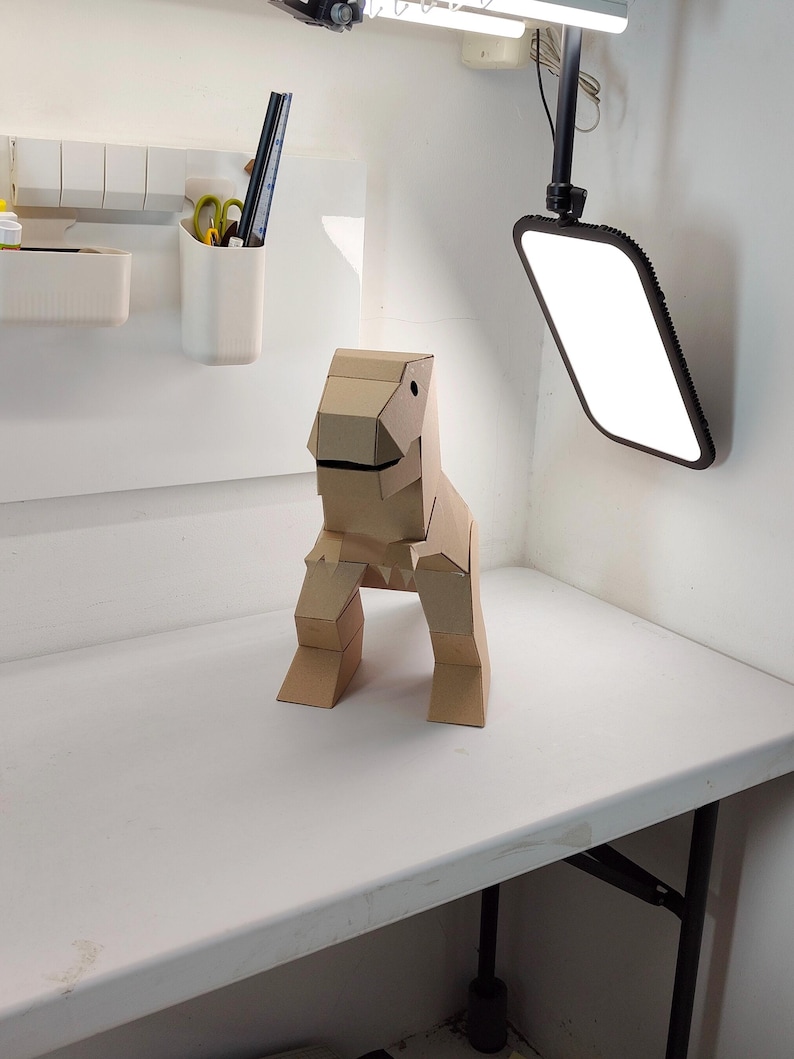 May include: A three-dimensional dinosaur figure constructed from light brown cardboard. The sculpture features a blocky, geometric design with a distinct head, a wide mouth, two short front limbs, and two sturdy back legs, all assembled from flat panels.