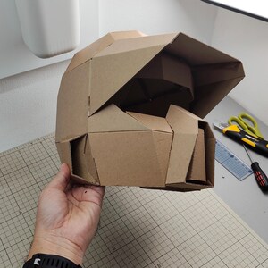 Op de afbeelding: Een geometrische helm gemaakt van bruin karton, met een gefacetteerd ontwerp met een opvallend vizier en een gestructureerde kinbeschermer. De helm is samengesteld uit meerdere hoekige panelen, wat zorgt voor een stijve, polygonale uitstraling.