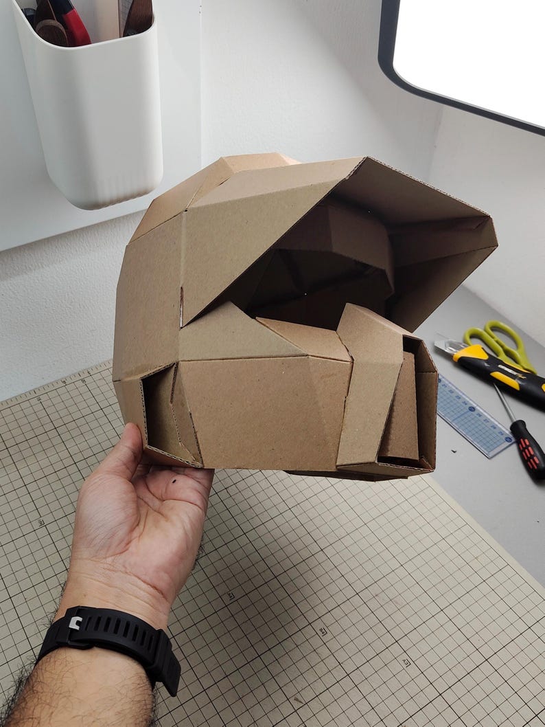 Op de afbeelding: Een geometrische helm gemaakt van bruin karton, met een gefacetteerd ontwerp met een opvallend vizier en een gestructureerde kinbeschermer. De helm is samengesteld uit meerdere hoekige panelen, wat zorgt voor een stijve, polygonale uitstraling.