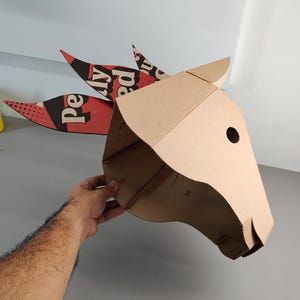 Puede incluir: Una m&aacute;scara de cabeza de animal hecha de cart&oacute;n marr&oacute;n con una oreja puntiaguda y tres accesorios decorativos en forma de cresta. Las crestas tienen patrones gr&aacute;ficos en rojo, negro y blanco con el texto "Pe" y "ly ed" en una fuente negrita.