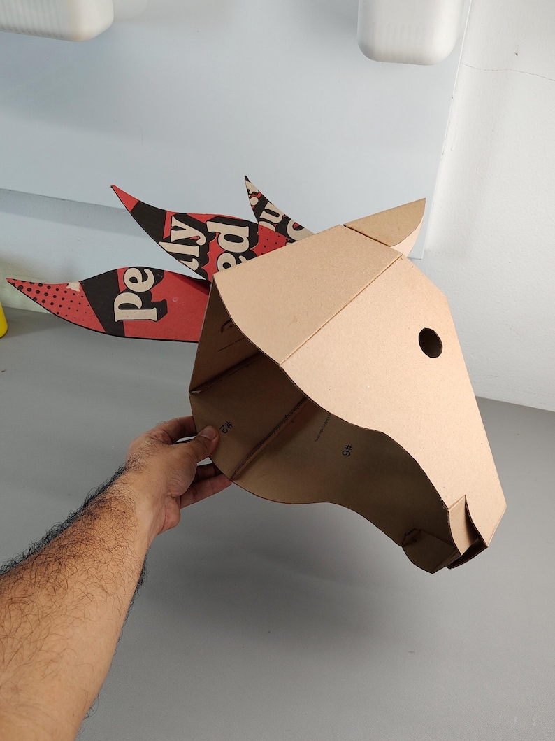 Puede incluir: Una m&aacute;scara de cabeza de animal hecha de cart&oacute;n marr&oacute;n con una oreja puntiaguda y tres accesorios decorativos en forma de cresta. Las crestas tienen patrones gr&aacute;ficos en rojo, negro y blanco con el texto "Pe" y "ly ed" en una fuente negrita.