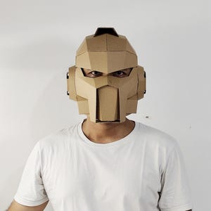 Könnte beinhalten: Eine Person trägt einen braunen Kartonhelm mit geometrischem Design. Der Helm hat eine rechteckige Frontplatte und kantige Merkmale. Die Person trägt ein weißes T-Shirt vor weißem Hintergrund.
