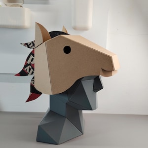 Puede incluir: Una m&aacute;scara de cabeza de caballo geom&eacute;trica de cart&oacute;n color canela, con un ojo circular negro y una crin hecha de papel estampado en negro, blanco y rojo, montada sobre un busto gris facetado con un perfil y estructura angulares.