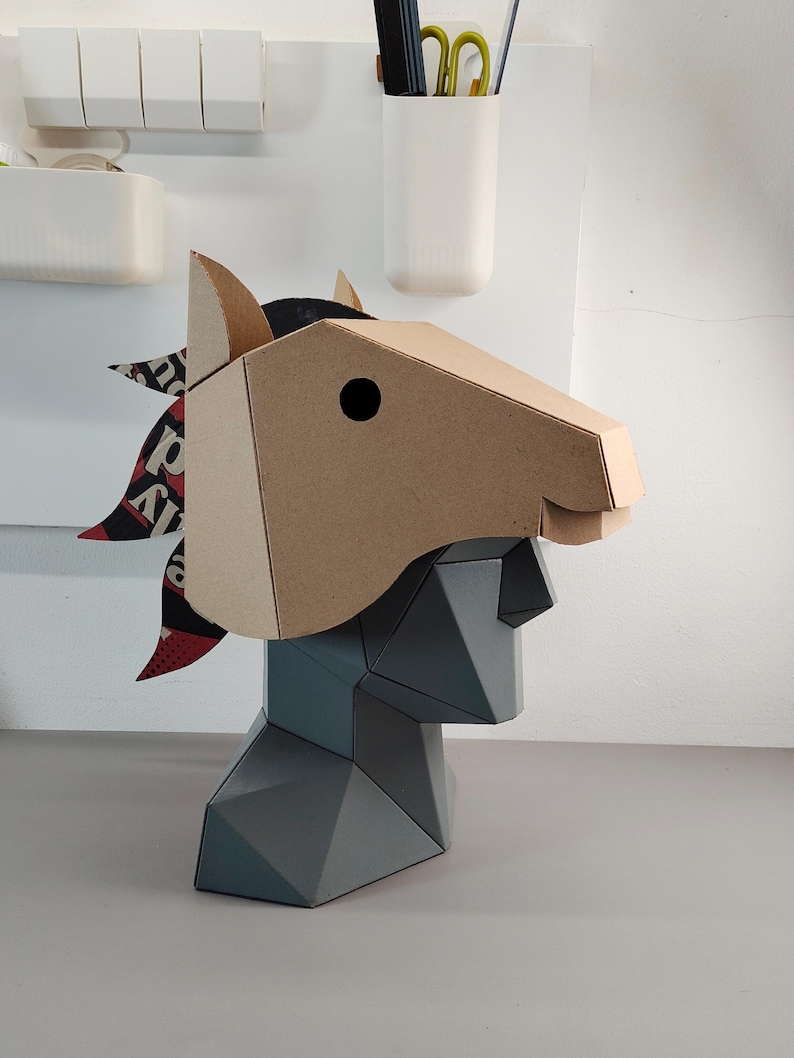 Puede incluir: Una m&aacute;scara de cabeza de caballo geom&eacute;trica de cart&oacute;n color canela, con un ojo circular negro y una crin hecha de papel estampado en negro, blanco y rojo, montada sobre un busto gris facetado con un perfil y estructura angulares.