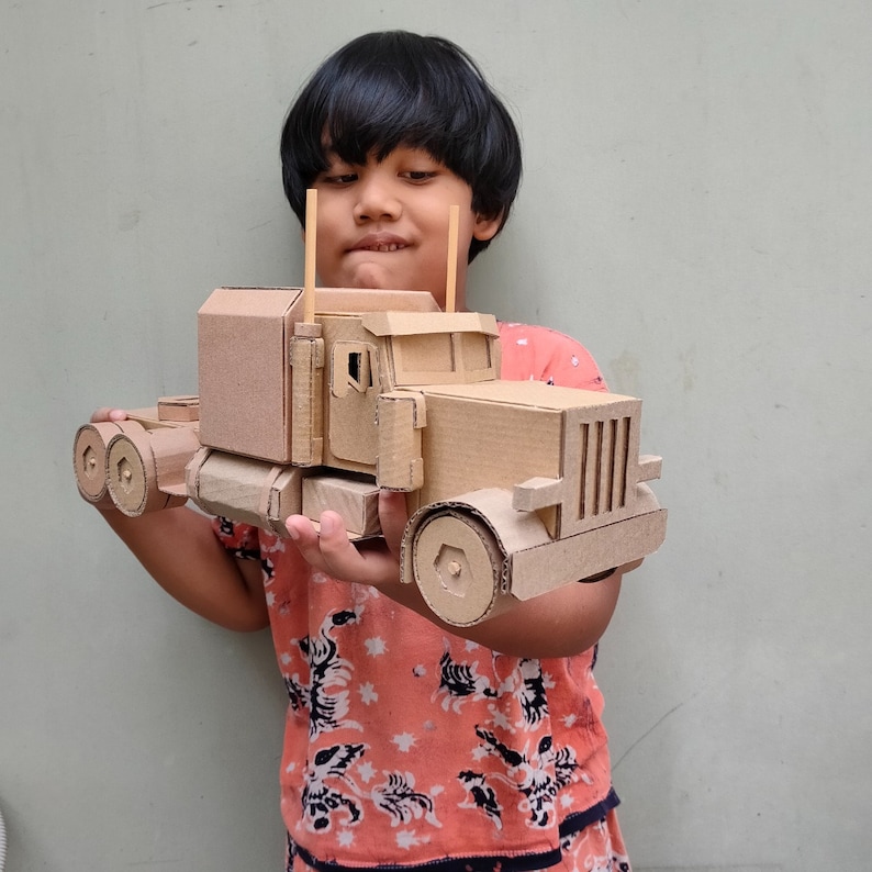 Pode incluir: Uma pessoa jovem segura um modelo de caminh&atilde;o de plataforma em papel&atilde;o. O caminh&atilde;o &eacute; marrom e tem uma cabine e um reboque detalhados. A pessoa est&aacute; vestindo uma camisa de estampa rosa e branca.