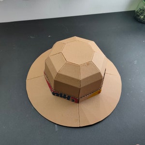Op de afbeelding: Een bruine kartonnen hoed met een geometrisch ontwerp. De hoed is gemaakt van meerdere stukken karton die aan elkaar zijn gelijmd. De hoed heeft een brede rand en een hoge kroon.
