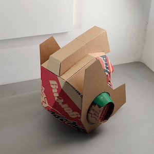 May include: A three-dimensional sculpture constructed from brown cardboard packaging. The structure features red and white printed branding with the text Indomie Goreng visible. A cylindrical green cardboard tube protrudes from the side.