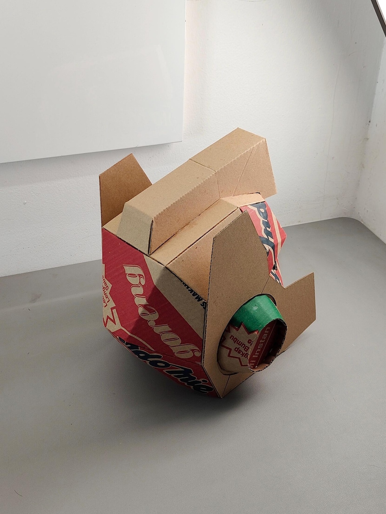 May include: A three-dimensional sculpture constructed from brown cardboard packaging. The structure features red and white printed branding with the text Indomie Goreng visible. A cylindrical green cardboard tube protrudes from the side.