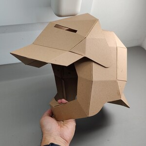 May include: A brown cardboard helmet with a visor and geometric design. The helmet is held by a hand, showcasing its three-dimensional structure and angular construction. The helmet's color is a natural, light brown.