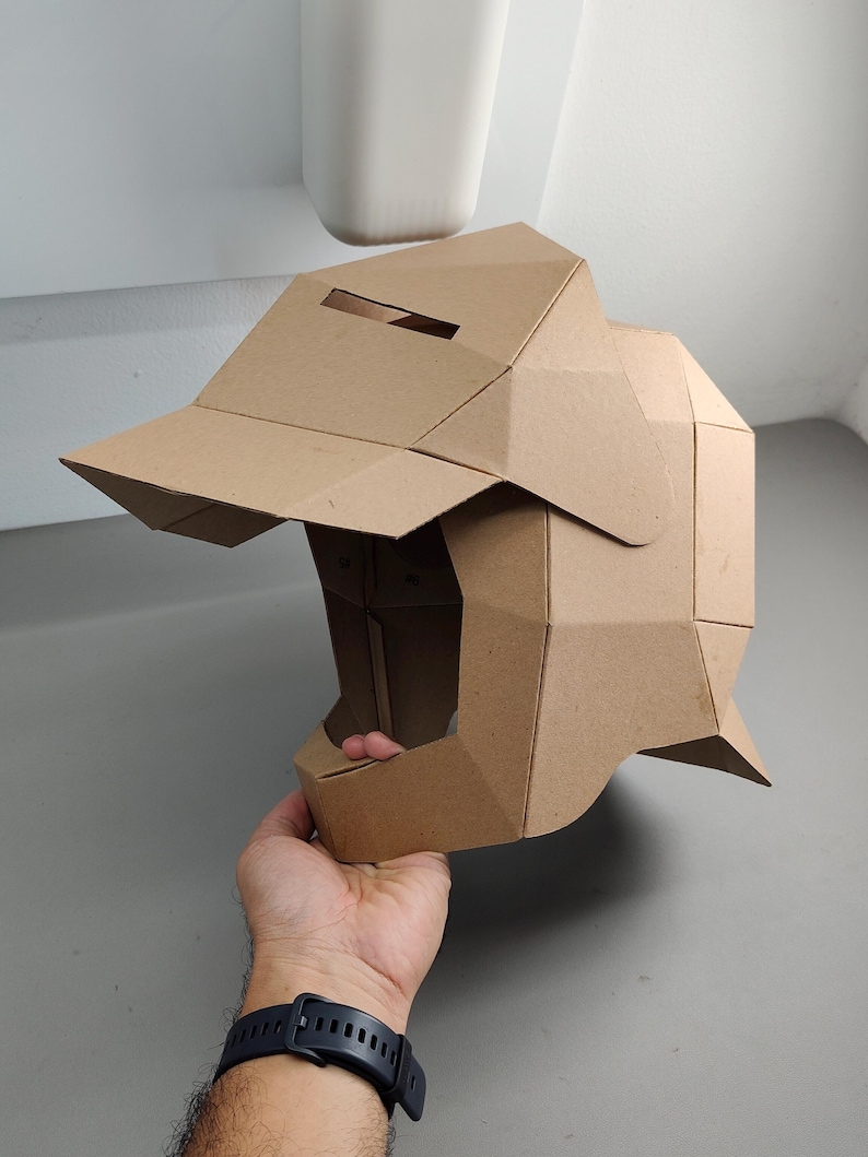 May include: A brown cardboard helmet with a visor and geometric design. The helmet is held by a hand, showcasing its three-dimensional structure and angular construction. The helmet's color is a natural, light brown.