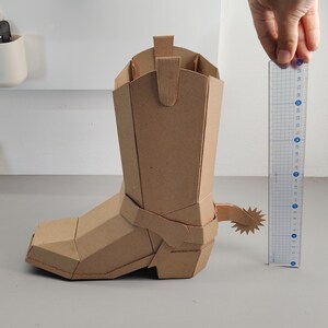 Puede incluir: Un modelo de bota vaquera de cart&oacute;n marr&oacute;n con una espuela en forma de estrella en el tal&oacute;n. Una regla de pl&aacute;stico transparente est&aacute; colocada junto a la bota, mostrando una altura de aproximadamente 20 cm desde la base hasta la parte superior.