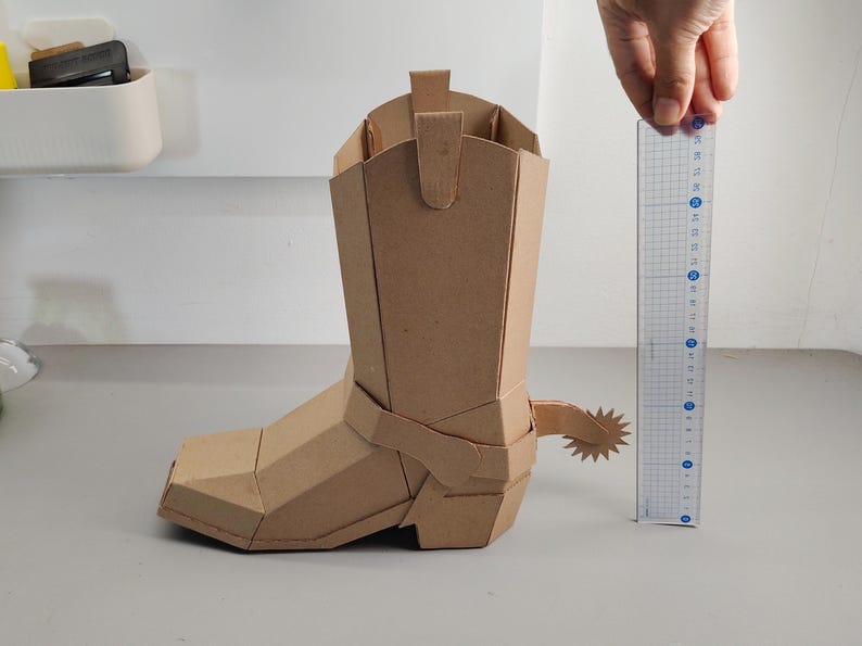 Puede incluir: Un modelo de bota vaquera de cart&oacute;n marr&oacute;n con una espuela en forma de estrella en el tal&oacute;n. Una regla de pl&aacute;stico transparente est&aacute; colocada junto a la bota, mostrando una altura de aproximadamente 20 cm desde la base hasta la parte superior.