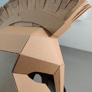 May include: A brown cardboard helmet featuring a geometric, angular design with a prominent crest on top. The helmet is constructed from multiple flat, interlocking panels of corrugated cardboard, showcasing a distinct layered texture.