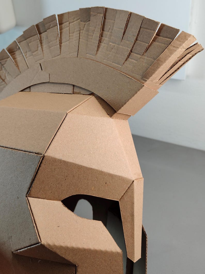 May include: A brown cardboard helmet featuring a geometric, angular design with a prominent crest on top. The helmet is constructed from multiple flat, interlocking panels of corrugated cardboard, showcasing a distinct layered texture.