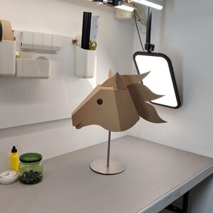 Puede incluir: Una escultura geom&eacute;trica de cabeza de caballo construida con paneles de cart&oacute;n beige planos. La pieza presenta orejas puntiagudas y una crin estilizada, montada sobre una varilla met&aacute;lica plateada vertical unida a una base circular de metal plateado.