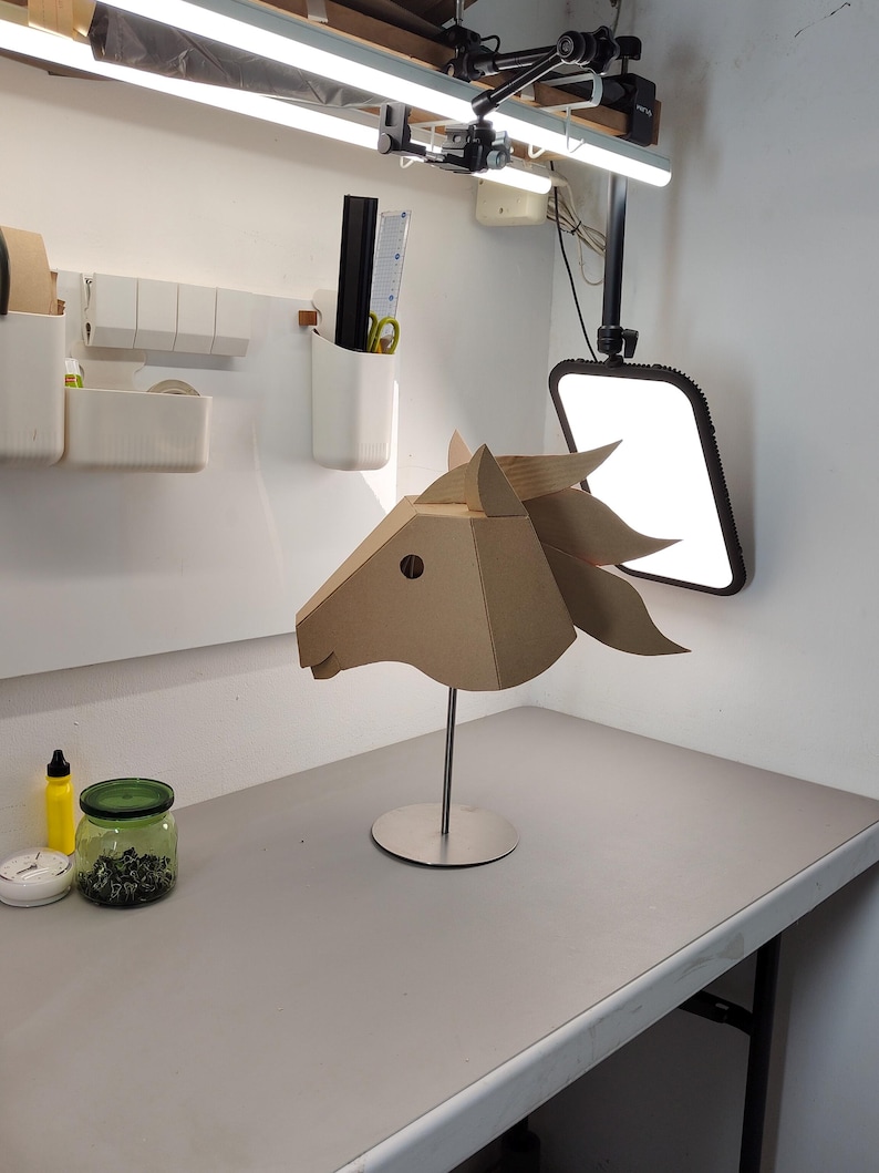 Puede incluir: Una escultura geom&eacute;trica de cabeza de caballo construida con paneles de cart&oacute;n beige planos. La pieza presenta orejas puntiagudas y una crin estilizada, montada sobre una varilla met&aacute;lica plateada vertical unida a una base circular de metal plateado.