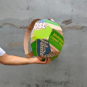 K&ouml;nnte beinhalten: Ein gr&uuml;ner, gelber und blauer Rucksack aus Pappe, hergestellt aus recycelten Lebensmittelverpackungen. Der Rucksack hat ein einzigartiges geometrisches Design und tr&auml;gt den Text "La Fonte" und "Makaroni Cepat Saji Quick Serve Macaroni" auf der Vorderseite.