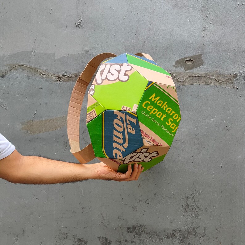 K&ouml;nnte beinhalten: Ein gr&uuml;ner, gelber und blauer Rucksack aus Pappe, hergestellt aus recycelten Lebensmittelverpackungen. Der Rucksack hat ein einzigartiges geometrisches Design und tr&auml;gt den Text "La Fonte" und "Makaroni Cepat Saji Quick Serve Macaroni" auf der Vorderseite.