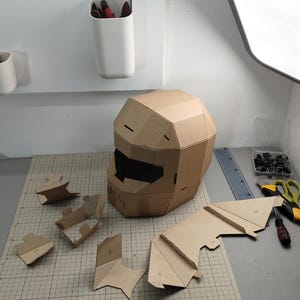 Op de afbeelding: Een bruine kartonnen helm met een geometrisch, gefacetteerd ontwerp en een donkere rechthoekige vizieropening. Verschillende kleinere, niet-gemonteerde kartonnen onderdelen met zichtbare cijfermarkeringen liggen op een snijmat.