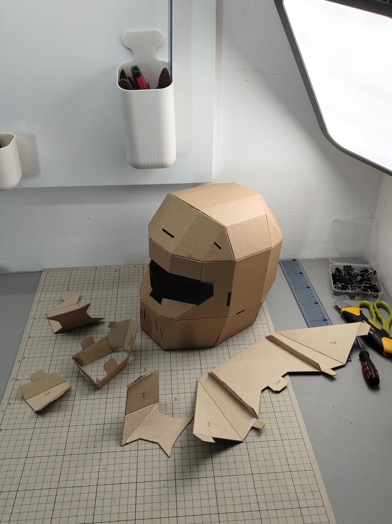 Op de afbeelding: Een bruine kartonnen helm met een geometrisch, gefacetteerd ontwerp en een donkere rechthoekige vizieropening. Verschillende kleinere, niet-gemonteerde kartonnen onderdelen met zichtbare cijfermarkeringen liggen op een snijmat.