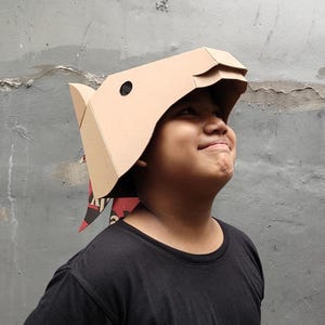 Puede incluir: Una m&aacute;scara de cart&oacute;n color canela con forma de cabeza de jabal&iacute; y dise&ntilde;o geom&eacute;trico angular. La m&aacute;scara presenta un recorte circular negro para el ojo y una secci&oacute;n de hocico. Una pieza de tela con estampado rojo y negro cuelga detr&aacute;s.