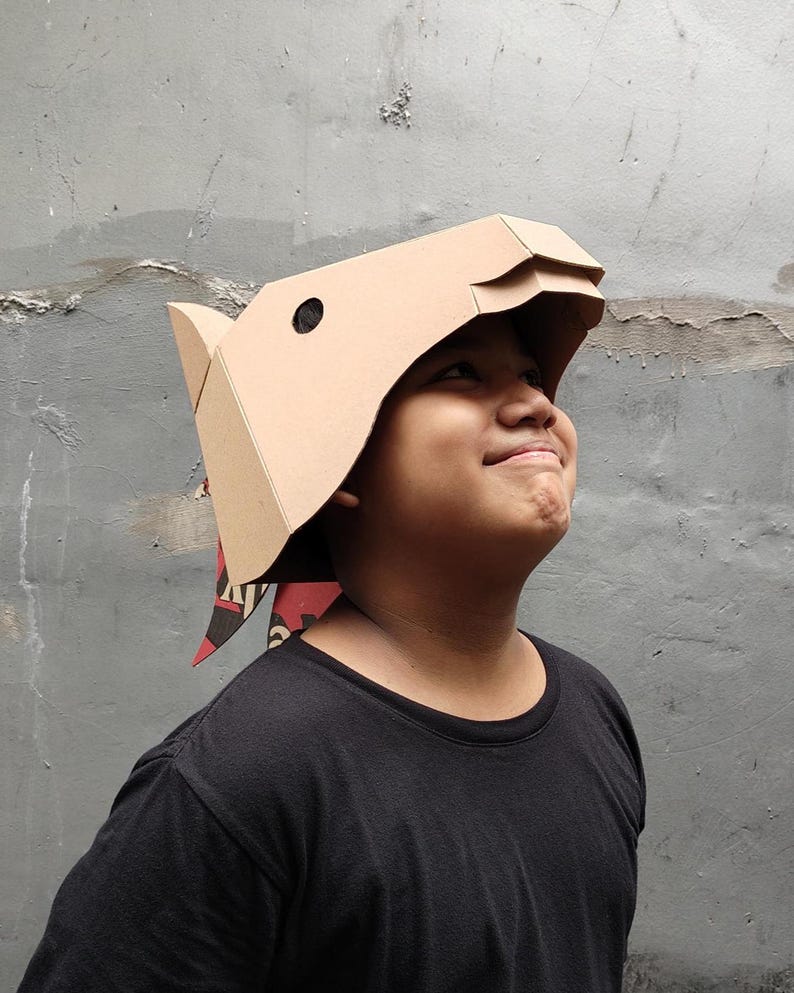 Puede incluir: Una m&aacute;scara de cart&oacute;n color canela con forma de cabeza de jabal&iacute; y dise&ntilde;o geom&eacute;trico angular. La m&aacute;scara presenta un recorte circular negro para el ojo y una secci&oacute;n de hocico. Una pieza de tela con estampado rojo y negro cuelga detr&aacute;s.