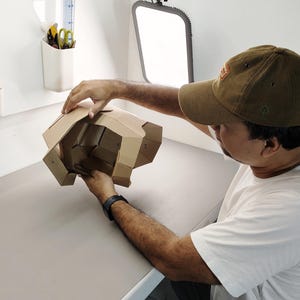 May include: A person holding a partially assembled cardboard structure. The structure is made of brown corrugated cardboard and has multiple folded sections. The person is wearing a brown cap and a white t-shirt, working in a well-lit studio setting.