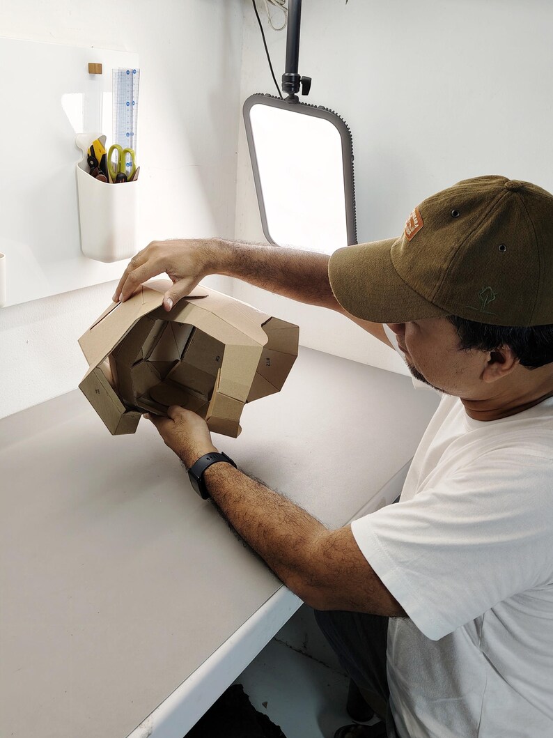 May include: A person holding a partially assembled cardboard structure. The structure is made of brown corrugated cardboard and has multiple folded sections. The person is wearing a brown cap and a white t-shirt, working in a well-lit studio setting.