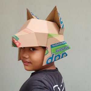 May include: A handmade cardboard helmet with pointed ears, constructed from repurposed cardboard boxes. The helmet features geometric shapes and visible text from the original packaging, showcasing a creative and resourceful design. The child is wearing a black shirt.