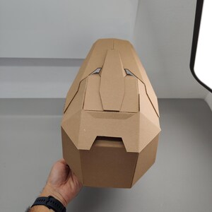May include: A geometric helmet constructed from light brown cardboard, featuring a faceted, angular design with a central vertical ridge and symmetrical side panels. The structure is composed of multiple interlocking pieces of tan paperboard.