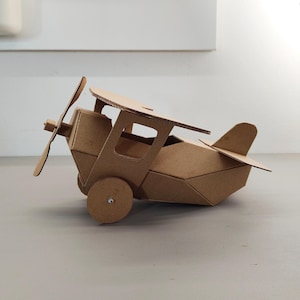 May include: A model aeroplane constructed from brown corrugated cardboard. The biplane features a front-mounted propeller, a cockpit with open side windows, two circular wheels on the sides, and a tail section with a horizontal stabiliser.