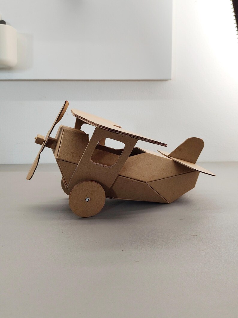 May include: A model aeroplane constructed from brown corrugated cardboard. The biplane features a front-mounted propeller, a cockpit with open side windows, two circular wheels on the sides, and a tail section with a horizontal stabiliser.