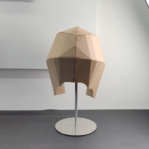 May include: A geometric helmet constructed from tan cardboard panels, featuring a faceted, angular design with a central seam. The helmet is mounted on a silver-coloured metal stand with a circular base, displaying a structured, multi-panelled form.