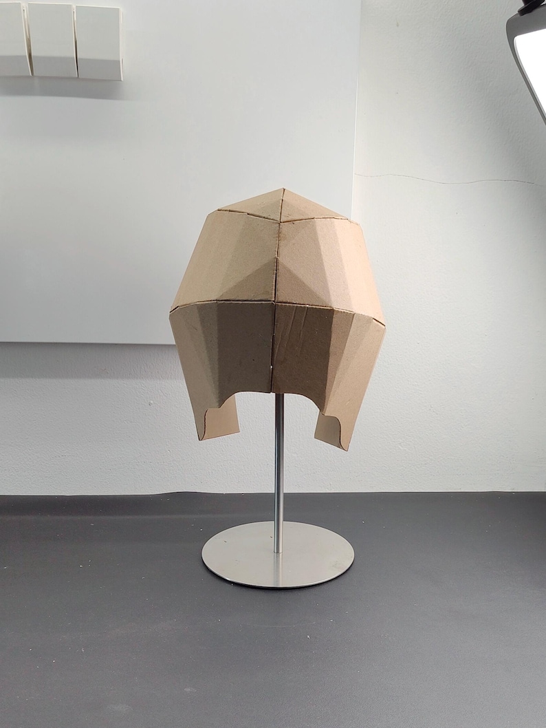 May include: A geometric helmet constructed from tan cardboard panels, featuring a faceted, angular design with a central seam. The helmet is mounted on a silver-coloured metal stand with a circular base, displaying a structured, multi-panelled form.