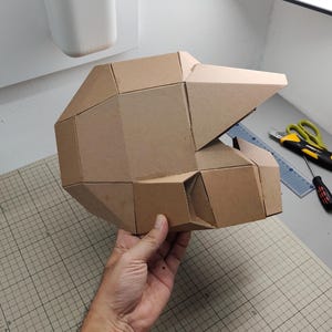 Op de afbeelding: Een geometrische helm opgebouwd uit bruine kartonnen panelen. De structuur heeft een gefacetteerd, hoekig ontwerp met een opvallend viziergedeelte en een holle binnenkant, wat een modulaire assemblage van platte, verbonden kartonnen stukken toont.