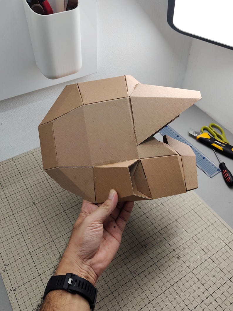 Op de afbeelding: Een geometrische helm opgebouwd uit bruine kartonnen panelen. De structuur heeft een gefacetteerd, hoekig ontwerp met een opvallend viziergedeelte en een holle binnenkant, wat een modulaire assemblage van platte, verbonden kartonnen stukken toont.