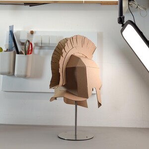 May include: A tan cardboard Roman-style helmet featuring a tall, segmented crest and a protective face guard. The helmet is mounted on a silver-colored metal stand with a circular base, showcasing a geometric construction made from folded cardboard.
