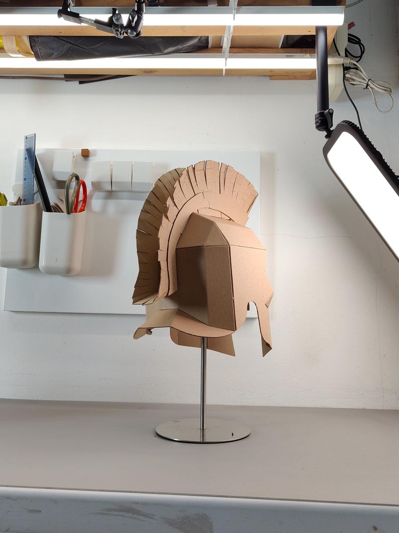May include: A tan cardboard Roman-style helmet featuring a tall, segmented crest and a protective face guard. The helmet is mounted on a silver-colored metal stand with a circular base, showcasing a geometric construction made from folded cardboard.