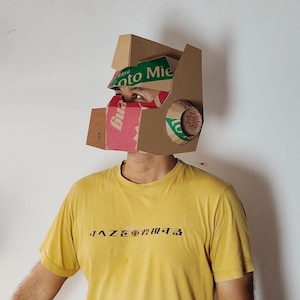 May include: A cardboard helmet constructed from recycled packaging materials, featuring brown corrugated cardboard panels with green and red printed text including the words 'Rasa' and 'Mie'. The helmet has a geometric design with a visor.
