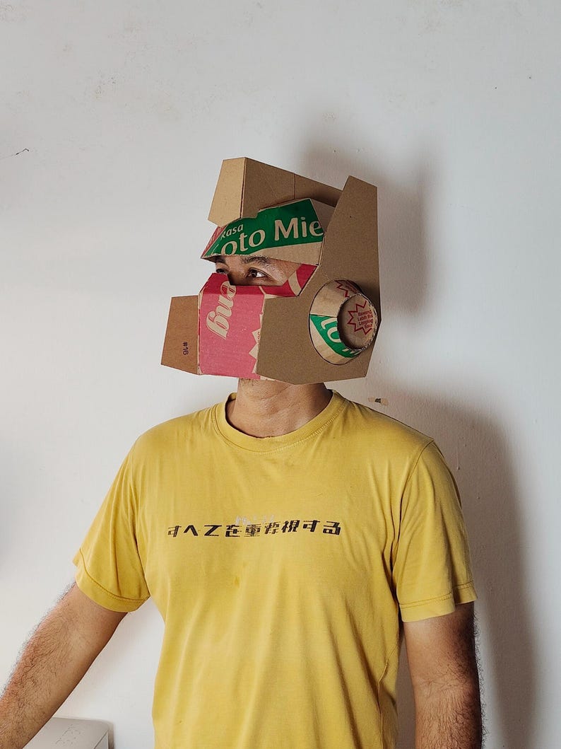 May include: A cardboard helmet constructed from recycled packaging materials, featuring brown corrugated cardboard panels with green and red printed text including the words 'Rasa' and 'Mie'. The helmet has a geometric design with a visor.
