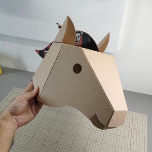 Puede incluir: Una m&aacute;scara de cabeza de caballo en tres dimensiones construida con cart&oacute;n corrugado color canela. La m&aacute;scara presenta un dise&ntilde;o geom&eacute;trico con un recorte circular para el ojo, orejas puntiagudas y una crin con patrones negros y rojos.
