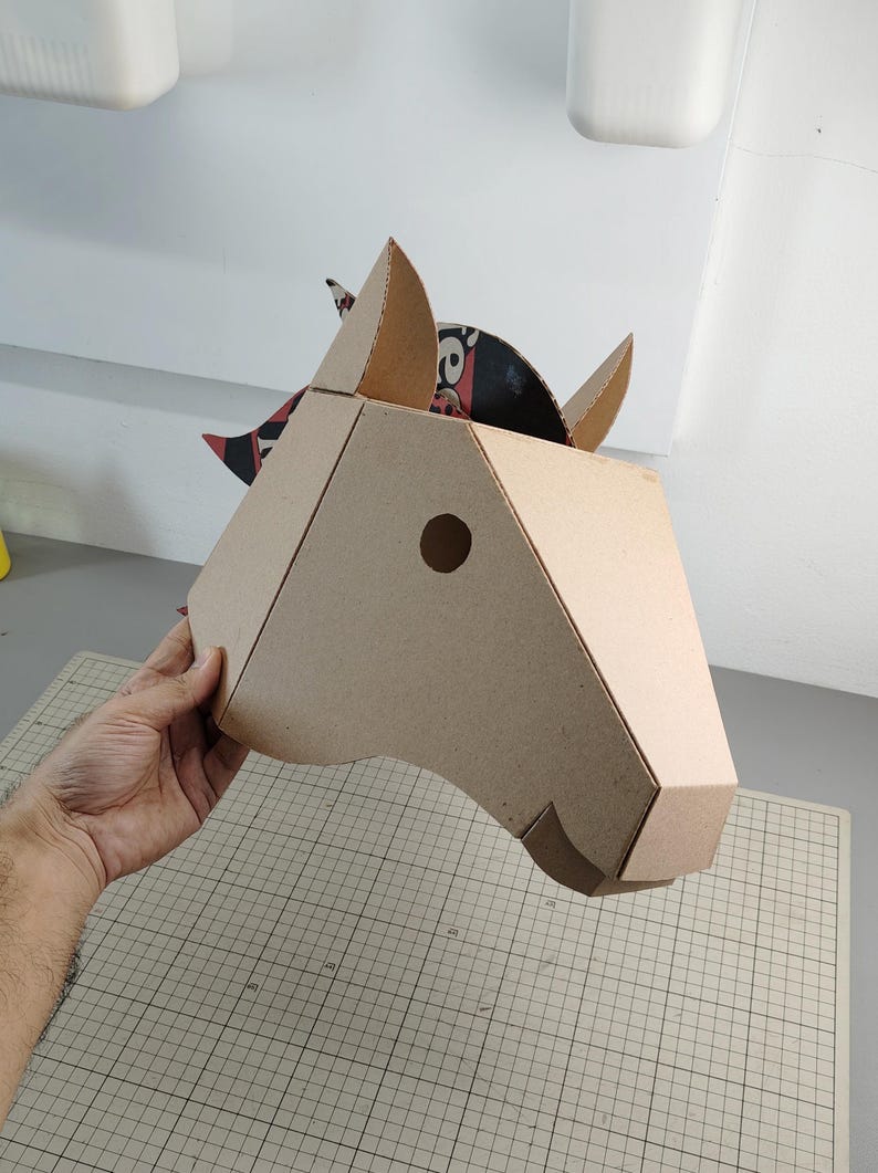 Puede incluir: Una m&aacute;scara de cabeza de caballo en tres dimensiones construida con cart&oacute;n corrugado color canela. La m&aacute;scara presenta un dise&ntilde;o geom&eacute;trico con un recorte circular para el ojo, orejas puntiagudas y una crin con patrones negros y rojos.