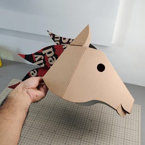 Puede incluir: Una m&aacute;scara de cabeza de caballo hecha de cart&oacute;n color canela con un dise&ntilde;o geom&eacute;trico. La crin est&aacute; hecha de papel estampado en rojo, negro y blanco con el texto visible "ly red" en fuente negrita. La m&aacute;scara tiene un recorte circular.