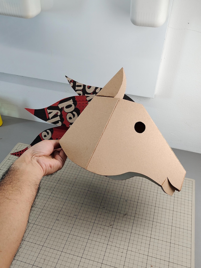 Puede incluir: Una m&aacute;scara de cabeza de caballo hecha de cart&oacute;n color canela con un dise&ntilde;o geom&eacute;trico. La crin est&aacute; hecha de papel estampado en rojo, negro y blanco con el texto visible "ly red" en fuente negrita. La m&aacute;scara tiene un recorte circular.
