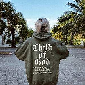 Pode incluir: Uma camisola de capuz verde com as palavras "Child of God" em letras brancas nas costas. O texto abaixo diz "And I will be a father to you, and you shall be sons and daughters to me, says the Lord Almighty, 2 Corinthians 6:18."