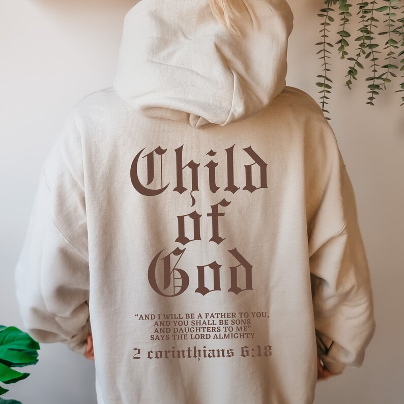 Pode incluir: Uma jaqueta de moletom bege com as palavras "Child of God" em letras g&oacute;ticas marrons. O texto "AND I WILL BE A FATHER TO YOU, AND YOU SHALL BE SONS AND DAUGHTERS TO ME" SAYS THE LORD ALMIGHTY 2 Corinthians 6:18" est&aacute; impresso abaixo do texto principal.