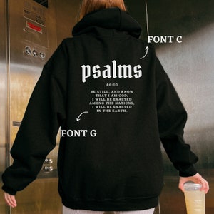 K&ouml;nnte beinhalten: Schwarzer Hoodie mit einem wei&szlig;en Aufdruck des Wortes "psalms" und einem Bibelvers aus Psalm 46:10. Der Text lautet "Seid still und erkennt, dass ich Gott bin; ich werde erh&ouml;ht werden unter den Nationen, ich werde erh&ouml;ht werden auf der Erde."