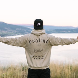 Puede incluir: Una sudadera con capucha beige con las palabras "psalms" y "LXII:11" impresas en la espalda. El texto contin&uacute;a con la cita "Truly he is my rock and my salvation; he is my fortress, I will never be shaken".